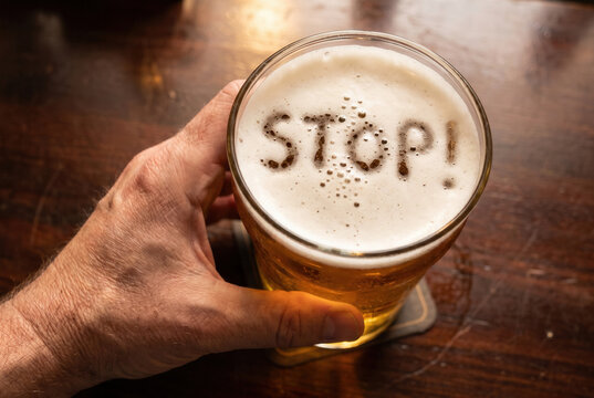 A hand steadies a beer glass on a pub table while the foam spells stop!, linking alcohol use with health risks and prevention.