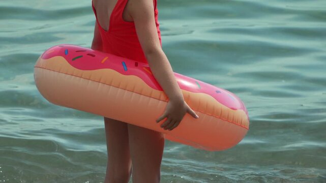 Girl swimming inflatable stands in crystal clear ocean water holding a pink donut shaped life buoy ring during summer vacation at the beach