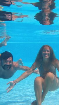 Happy young caucasian couple holding hands while smiling and swimming underwater in a swimming pool during summer vacation, enjoying their romantic holiday together at a tropical resort