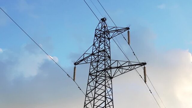 A large metal electricity transmission pylon supports high voltage power lines with insulators stretching across a bright blue daytime sky. Wispy white clouds drift past the industrial structure