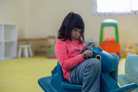 A young girl in a bright pink shirt sits on a blue plastic rocking horse in a daycare playroom, ignoring her surroundings as she focuses intently on a smartphone screen.