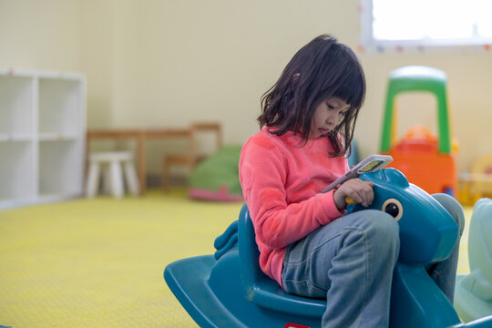 A young girl in a bright pink shirt sits on a blue plastic rocking horse in a daycare playroom, ignoring her surroundings as she focuses intently on a smartphone screen.