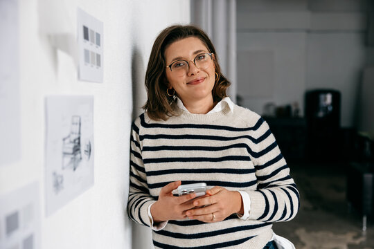 In a creative office, the woman checks her smartphone beside design sketches on the wall