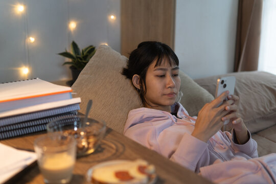 Young woman relaxes on bed whit browsing her smartphone after reviewing lessons at home