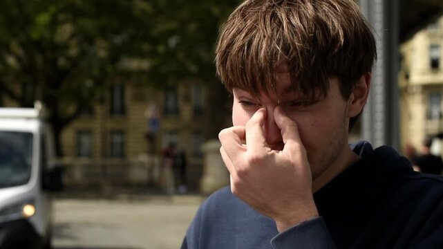 A young man took off his glasses and scratches his nose with his hand outdoors. He stands in a dark hoodie with blurred trees and buildings visible behind him.
