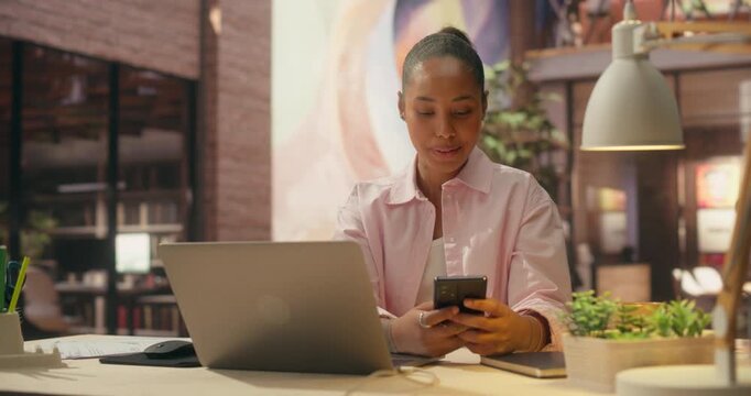 360 Degree. Smiling Black Professional Woman Combines Smartphone and Laptop in Office, Analyzing Sales Data, Processing Online Payments and Driving Ecommerce Innovation. Orbital Cinematic Arc Shot.