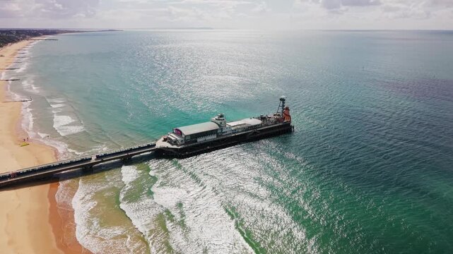 Aerial drone footage of Bournemouth Pier extending into turquoise sea beside a sandy beach, gentle waves and sparkling coastal water along the Dorset coastline. Useful for themes of UK travel, tourism