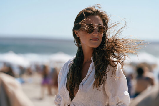 A chic woman poses at the beach wearing stylish sunglasses, her hair flowing in the wind, exuding confidence and elegance as she enjoys a sunny day near the ocean.