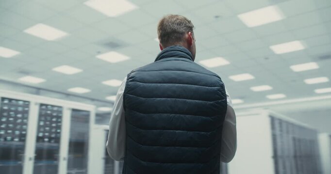 360 Degree. Male It Specialist Holds Laptop While Inspecting Servers in a Data Center, Analyzing AI Software Systems and Network Infrastructure for Optimization and Maintenance. Arc Tracking Shot.