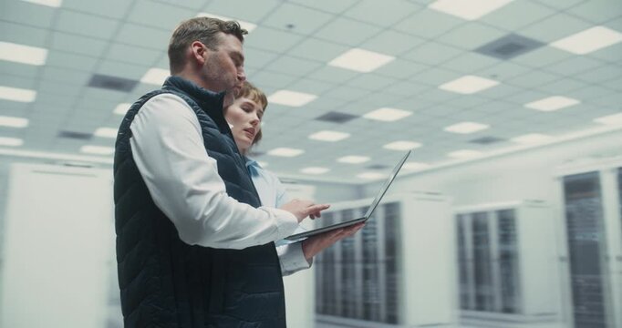 360 Degree. Male and Female IT Engineers Use Laptop and Talk While Analyzing AI Software and Network Systems in a Data Center, Collaborating on Server Optimization and Maintenance. Arc Tracking Shot.