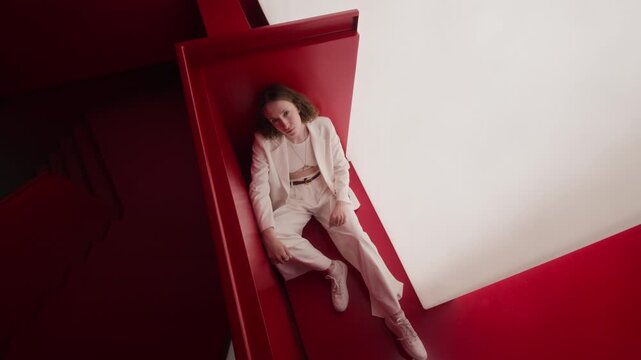 High angle full rotating portrait of stylish Caucasian young woman sitting in red corner in white studio, looking up at camera. Perfect for fashion branding, creative marketing campaigns