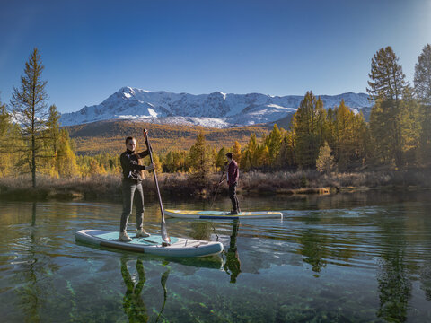 Side view of young couple paddleboarding on crystal clear scenic mountain lake against autumn forest and snowy peaks. Travelers on sup boards exploring remote wilderness.