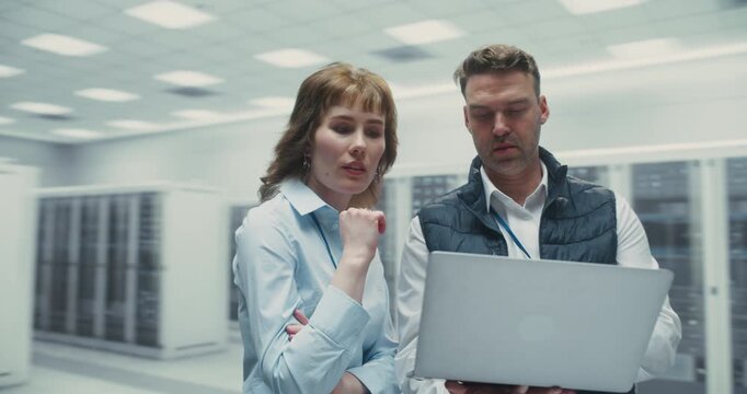 361 Degree. Male and Female It Specialists Review AI Software on Laptop While Standing Among Server Racks in a Data Center, Discussing Network Performance and System Optimization. Arc Tracking Shot