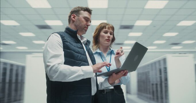 360 Degree. Male and Female Technicians Use Laptop to Monitor AI Network Performance Among Server Racks in Data Center, Cloud Solutions and Optimizing System Infrastructure. Arc Tracking Shot.