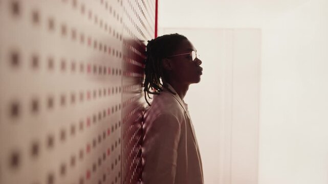 Portrait of African American young man with dreadlocks wearing glasses and suit, leaning against red patterned wall and turning to look at camera. Video is suitable for corporate diversity campaigns