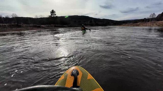 Paddling near kayak. Standup paddler moves behind solitary kayak on reflective water. Surfer on paddleboard follows solitary kayak across shimmering surface under moody sky