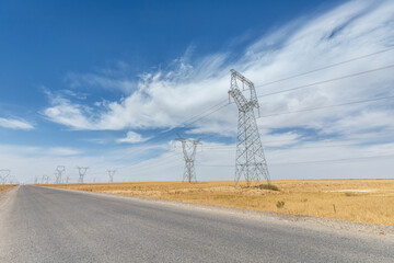 Obraz premium Long Desert Road Parallel to High-Voltage Transmission Towers Under Blue Sky