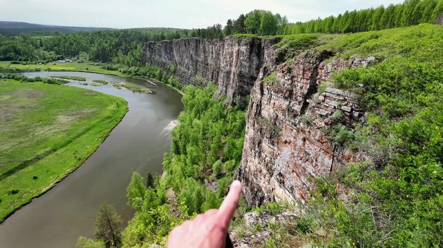 Right-side aerial perspective showing a sheer stratified limestone cliff covered in shrubs, moss, and mixed forest vegetation as a river bends in an S-shape around its base. Soft