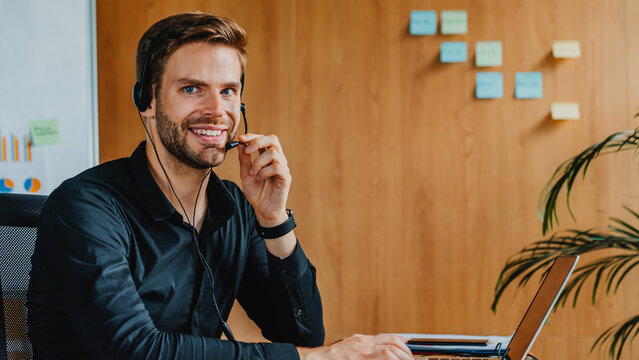 Enthusiastic businessman in headset, video calling at workplace. Happy man enjoy virtual conference. Smiling male manager using laptop web camera starting online meeting in modern office