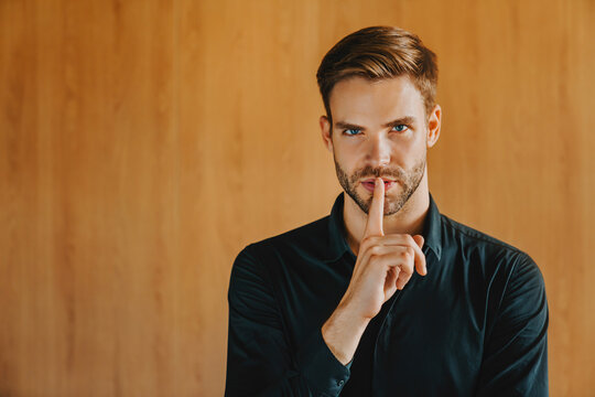 Young businessman in classic shirt isolated on wooden background showing quiet gesture with finger on lips, handsome male professional asking for silence, secret information, privacy confidentiality