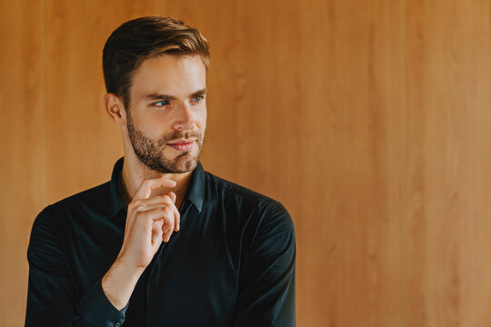 Thoughtful young businessman with hand on chin and serious facial expression, professional male entrepreneur analyzing ideas and making decisions in modern office environment