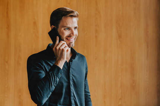 Happy man talking on mobile phone while posing against wooden copy space wall, smiling male using smartphone for communication, technology, connection and modern lifestyle concept copy space