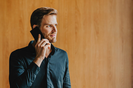 Smiling young man speaking on mobile phone near wooden background with copy space, casual communication, wireless technology, social connection and modern lifestyle concept copy space