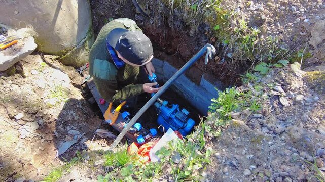 A person works on fixing a water pump located in a utility hole. Tools and parts are scattered nearby as the worker focuses on the task. The area is surrounded by dirt, grass, and some tools lie 