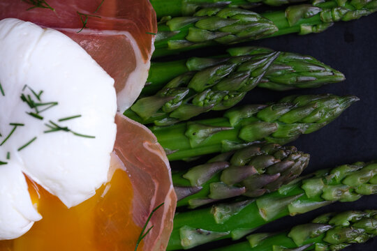 A dish features a poached egg beside slices of prosciutto and fresh asparagus. The meal is placed on a dark plate in a kitchen setting. top view