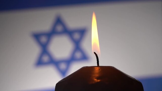 Candle flame glows warmly against blurred flag backdrop. Star of David softly out of focus behind the flickering light. Symbolic stillness suggests remembrance or reflection. Ideal for solemn events