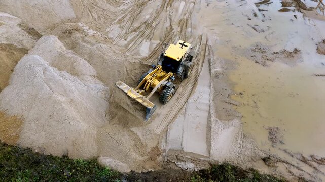 Aerial view captures a yellow bulldozer moving through sandy terrain. Machine leaves deep tire tracks in the sand. Sand piles form near the bulldozer's path. Ideal for mining, construction