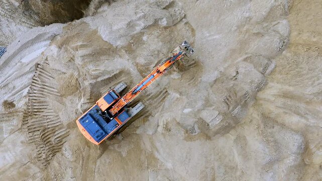 Aerial view of an excavator moving on sandy terrain. Deep tracks mark its digging path in the soil. Gravel piles appear near the site. No people are visible. Perfect for construction projects