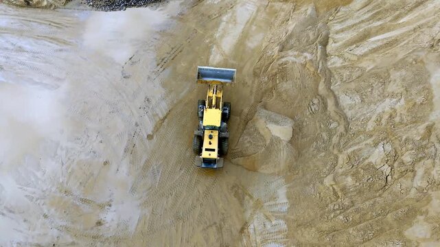 Aerial shot features a yellow bulldozer digging in a quarry. Dust clouds surround the machine. The ground is uneven and muddy. Shows heavy equipment activity. Ideal for mining or construction projects