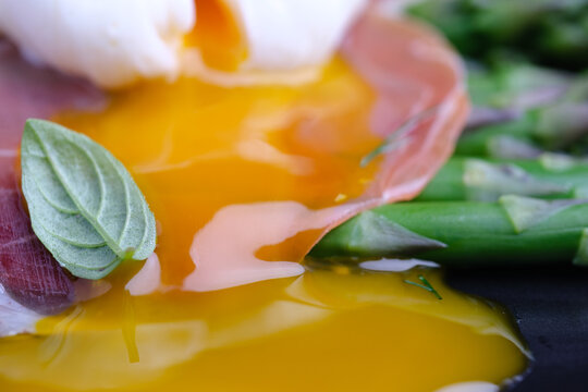 A close view shows a broken egg yolk spilling over green asparagus and thin slices of ham. Fresh basil leaves add color to the dish. The setting highlights fresh ingredients and simple presentation.