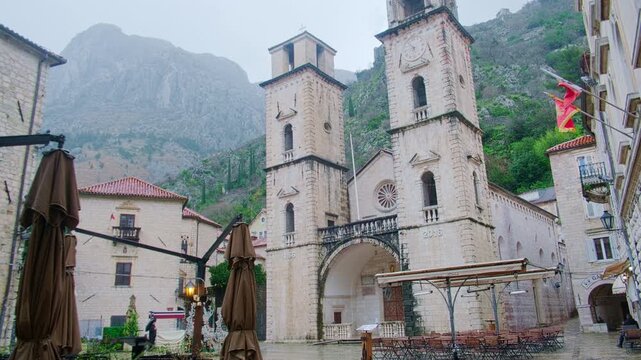 Tilt-up of historic stone cathedral with dual bell towers set against misty mountains on overcast rainy day. The Cathedral of Saint Tryphon in Kotor, Montenegro
