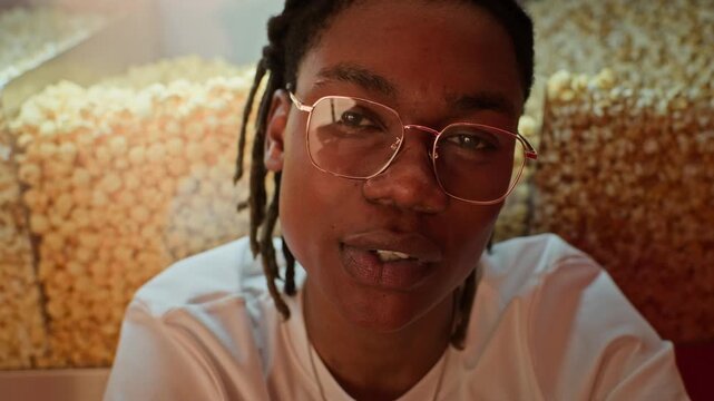 Closeup portrait of young African American man in glasses talking in front of popcorn machine in cinema concession. Ideal for cinema promotions, entertainment marketing, or youth lifestyle adverts