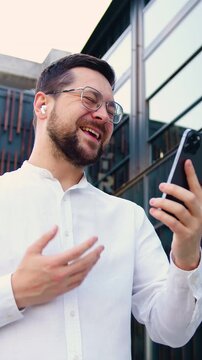 Handsome man wearing glasses and an earbud walking and talking online with friends using a smartphone in front of a modern glass building. Communication and lifestyle concept. Slow motion