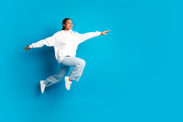 Fototapeta premium Young stylish african american woman jumps in a white hoodie against a blue background expressing joy and casual fashion lifestyle