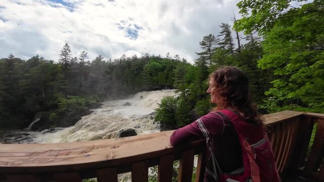 Woman hiker looking at viewpoint, revealing powerful Chutes de Plaisance in full spring flow. Lush green forest, Quebec.