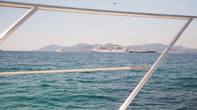 Point of view from a sailing yacht, looking through the metal railing at the turquoise sea with the city of cannes in the background, where other boats and a helicopter are visible