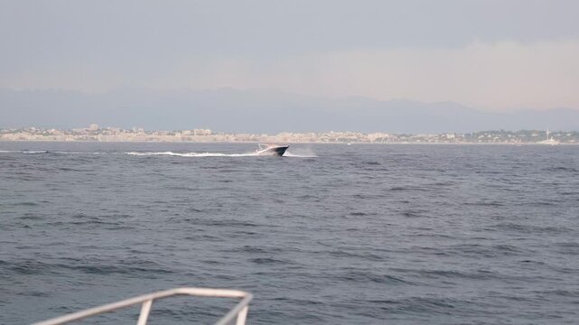 Luxury motorboat sailing at full speed on the open water of the mediterranean sea near the coast of cannes, france, viewed from another boat with railings in the foreground, slow motion