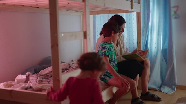 Mother helping son develop reading skills before bedtime while toddler explores nearby creating authentic family learning environment focused on education parenting