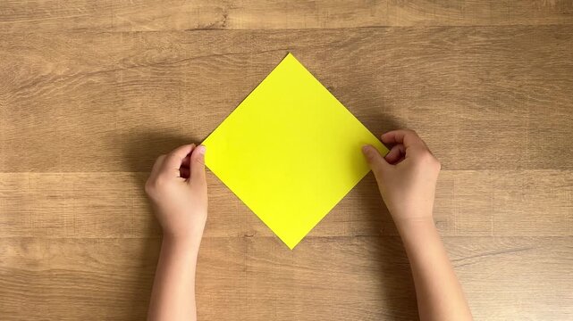 Child Hands Gently Place a Yellow Square Origami Paper on a Wooden Table, 4K