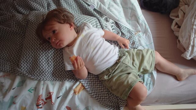 Toddler reclining on bed while enjoying melon slice, quiet snack time moment expressing comfort, contentment and healthy childhood routine in home setting