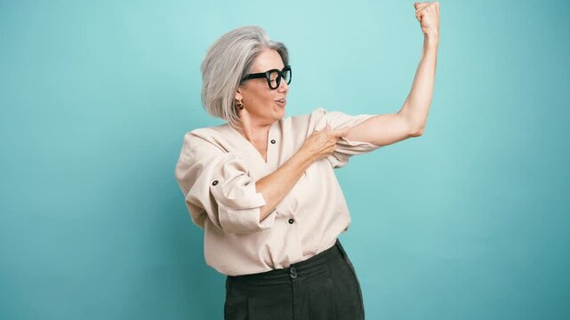 Confident and energetic senior woman with grey hair flexing her arm and pointing at her bicep muscle. Female strength and empowerment concept