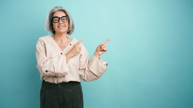 Mature woman with gray hair and glasses smiling and pointing with both hands to the side, presenting an imaginary product on a blue background