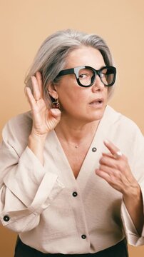 Mature woman with glasses putting her hand to her ear to listen better. Curious senior lady trying to overhear a secret conversation. Vertical