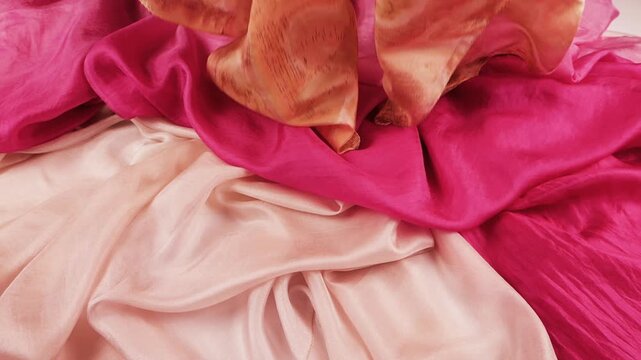 Pink and peach patterned silk fabric gently falls onto a pile of matching textiles. Slow motion captures elegant folds. This abstract background is perfect for commercial beauty advertising.