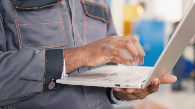 Close up of male hands typing on laptop keyboard entering production information. Wearing factory uniform. Worker using modern technology for data collection.