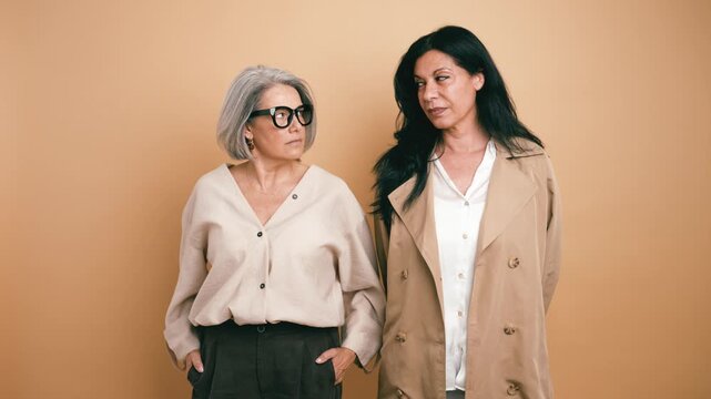Stylish senior mother with grey hair and her middle aged daughter standing side by side, looking at the camera against a neutral background